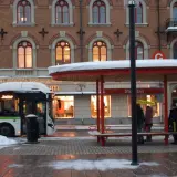 Snö faller vid en busshållplats där folk väntar. En buss med destination "tåg till Sundsvall" står parkerad.
