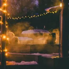 Två personer njuter av ett varmt bad utomhus i snön, upplysta av julbelysning. En mysig kväll på ett kallbadhus i Stockholm.