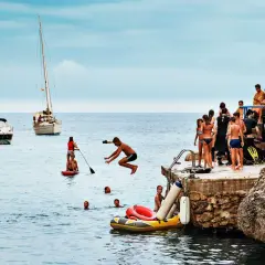 Barn hoppar i vattnet vid en klippig kust, en segelbåt och motorbåtar i bakgrunden. En perfekt dag för bad och lek vid Palma de Mallorca.