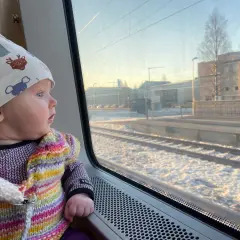 Litet barn i färgglad kofta och mössa med djurmotiv tittar ut genom tågfönstret. Snö på marken och byggnader syns utanför. Kanske är det ett nattåg till Bryssel?