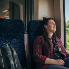 Ung kvinna med hörlurar njuter av utsikten från ett tåg. Hennes ryggsäck står redo för äventyr när hon ska backpacka Europa.