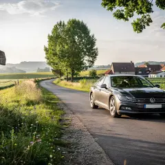 En mörkgrå Volkswagen kör längs en slingrande väg genom ett lantligt europeiskt landskap. En gammal vägvisare pekar mot en okänd destination, perfekt för en bila i Europa rutt.