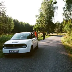 Vit elbil parkerad vid en skogsväg, redo för resan från Stockholm till Oslo. Två personer står vid sidan av vägen.