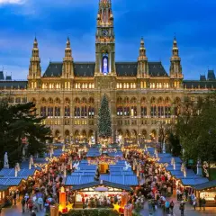 En livlig julmarknad i Europa framför ett upplyst stadshus. Träd med röda dekorationer och en stor julgran bidrar till den festliga stämningen.