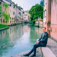 Man sitter vid en kanal i Treviso, Italien. Vatten reflekterar färgglada hus och gröna träd.