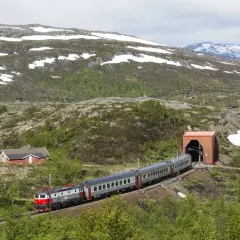 Tåg på väg mot tunneln, en del av sträckan Kiruna Narvik, med snö på bergssluttningarna och små röda hus.