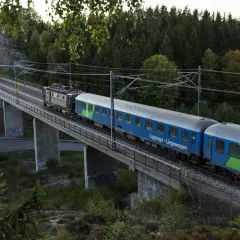Ett blått SJ-tåg, kanske ett nattåg Berlin, korsar en bro genom en skogig miljö.