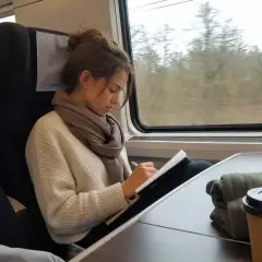 Ung kvinna skriver i en anteckningsbok på ett db tåg. Utanför fönstret syns träd.