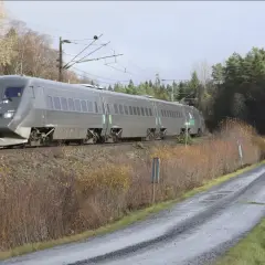 Grått SJ-tåg på väg mot Stockholm, passerar en slingrande grusväg och höstfärgad natur.