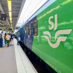 Folk väntar på ett grönt och blått tåg med SJ-loggan på perrongen. Kanske på väg till London?