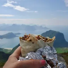 En hand håller en burrito med en fantastisk utsikt över Lofoten. En perfekt paus under vandringen.