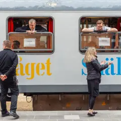 Män tittar ut genom fönstret på ett tåg med texten "Snälltåget" och destinationen Innsbruck. En kvinna gestikulerar mot tåget.