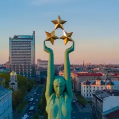 Frihetsmonumentet i Riga reser sig mot en solnedgångshimmel, med tre gyllene stjärnor högt upp. En båt syns i fjärran.