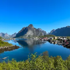 En klarblå himmel speglar sig i det lugna vattnet vid en idyllisk by. Majestätiska berg reser sig bakom de röda husen. En vacker lofoten sommarvy.