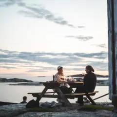 Två personer njuter av en middag vid ett picknickbord vid vattnet. En perfekt hemester i Stockholms skärgård.