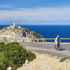 En cyklist njuter av en cykelsemester i Europa, med en fyr på en klippa vid det blå havet i bakgrunden.