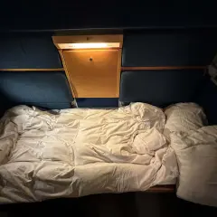 Ett bäddat nattågskupé med vita lakan och kuddar. En liten trähylla med lampa finns ovanför sängen.