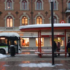 Snö faller vid en busshållplats där folk väntar. En buss med destination "tåg till Sundsvall" står parkerad.