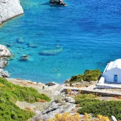 En vit kyrka med blå dörr vid en turkos vik på Amorgos, Grekland. Några badar i det klara vattnet.