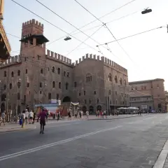 En solig dag i Bologna, Italien. En stor medeltida byggnad med torn dominerar torget, där människor promenerar och sitter vid uteserveringar.