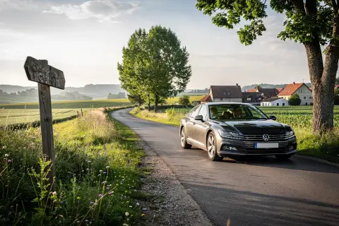 En mörkgrå Volkswagen kör längs en slingrande väg genom ett lantligt europeiskt landskap. En gammal vägvisare pekar mot en okänd destination, perfekt för en bila i Europa rutt.