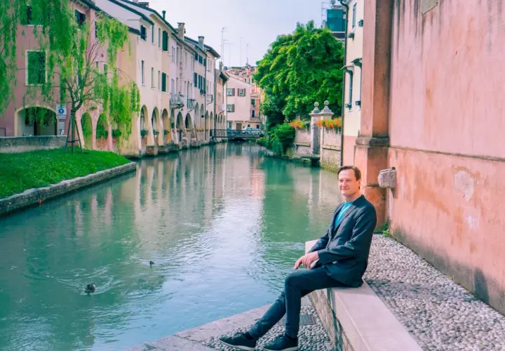 Man sitter vid en kanal i Treviso, Italien. Vatten reflekterar färgglada hus och gröna träd.