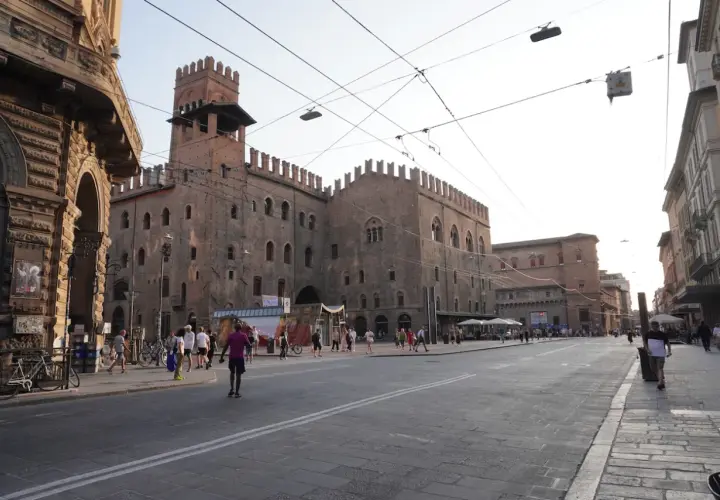 En solig dag i Bologna, Italien. En stor medeltida byggnad med torn dominerar torget, där människor promenerar och sitter vid uteserveringar.