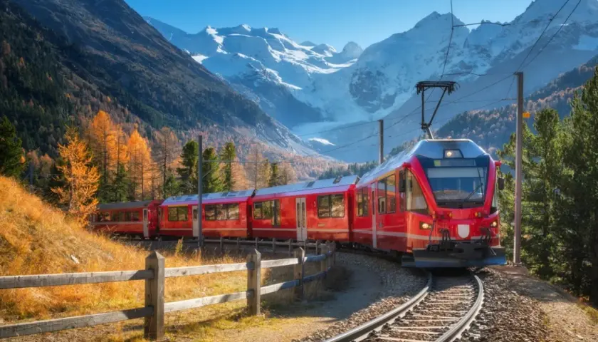 Rött tåg på Bernina Express-linjen, perfekt för Interrail, slingrar sig genom en höstfärgad dal med snöklädda berg i bakgrunden.