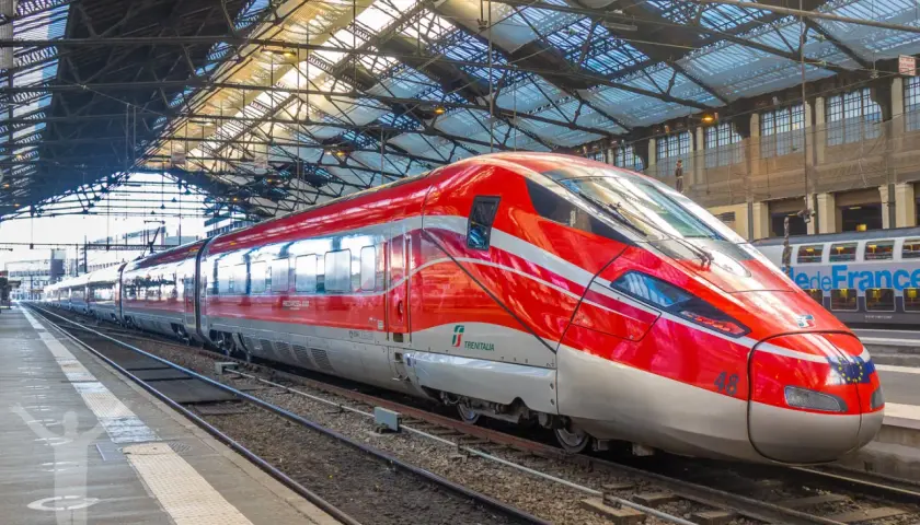 Ett rött snabbtåg i Frankrike, med namnet "Frecciarossa 1000", står på en station under ett glastak.