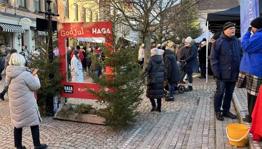 Människor njuter av en livlig julmarknad i Haga. En röd ram med texten "God Jul från HAGA" inbjuder till foton bland granar och stånd.