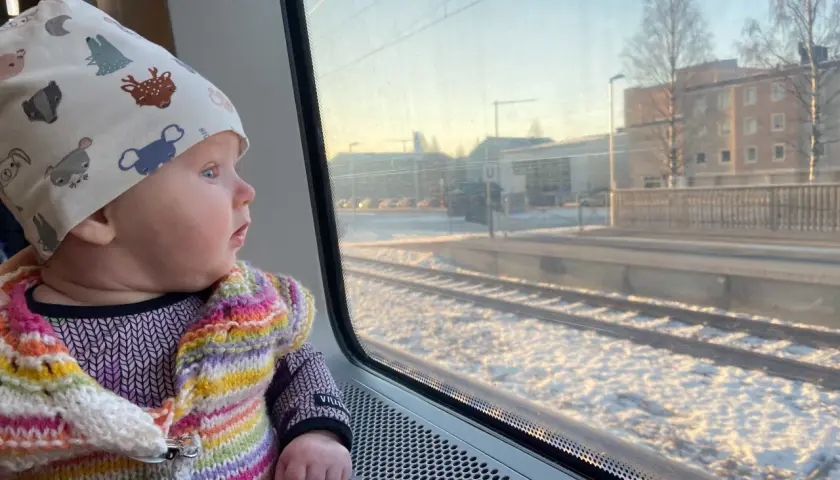Litet barn i färgglad kofta och mössa med djurmotiv tittar ut genom tågfönstret. Snö på marken och byggnader syns utanför. Kanske är det ett nattåg till Bryssel?