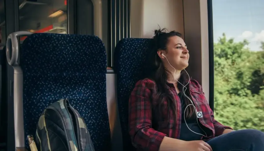 Ung kvinna med hörlurar njuter av utsikten från ett tåg. Hennes ryggsäck står redo för äventyr när hon ska backpacka Europa.