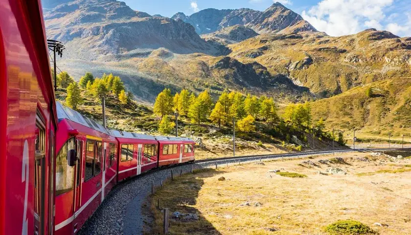 Rött tåg slingrar sig genom ett bergigt landskap med höstfärger. Ett perfekt sätt att uppleva Europa, funderar du på interrail pris?