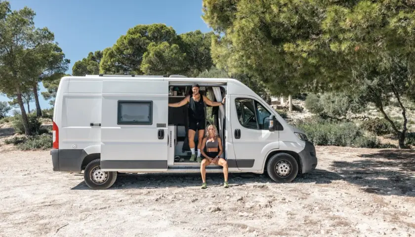 Ett par njuter av van life vid en solig plats med träd. Mannen står i dörröppningen till husbilen, kvinnan sitter på tröskeln.