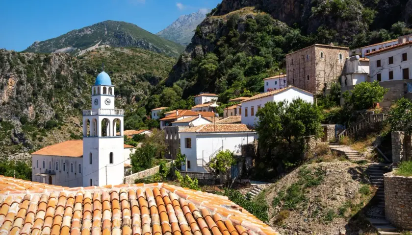 En vit kyrka med blå kupol reser sig i en bergig by. Kanske är detta en del av resan med tåg till Albanien, en vy av traditionell arkitektur mot en dramatisk bakgrund.