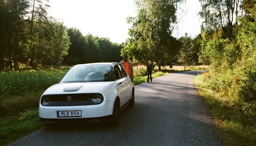 Vit elbil parkerad vid en skogsväg, redo för resan från Stockholm till Oslo. Två personer står vid sidan av vägen.