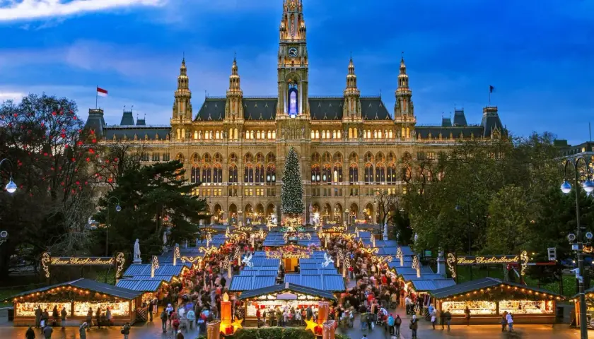 En livlig julmarknad i Europa framför ett upplyst stadshus. Träd med röda dekorationer och en stor julgran bidrar till den festliga stämningen.
