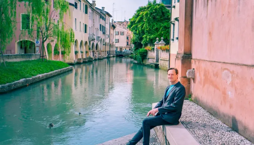 Man sitter vid en kanal i Treviso, Italien. Vatten reflekterar färgglada hus och gröna träd.