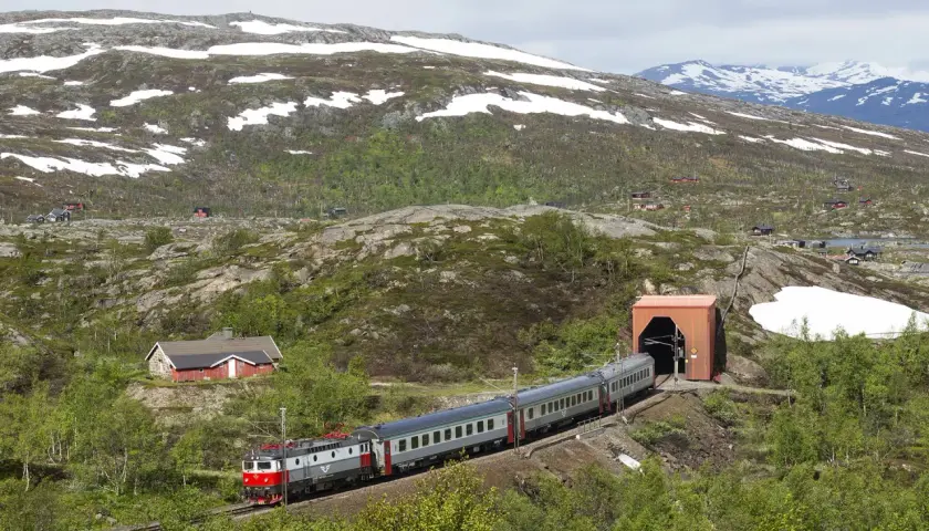 Tåg på väg mot tunneln, en del av sträckan Kiruna Narvik, med snö på bergssluttningarna och små röda hus.