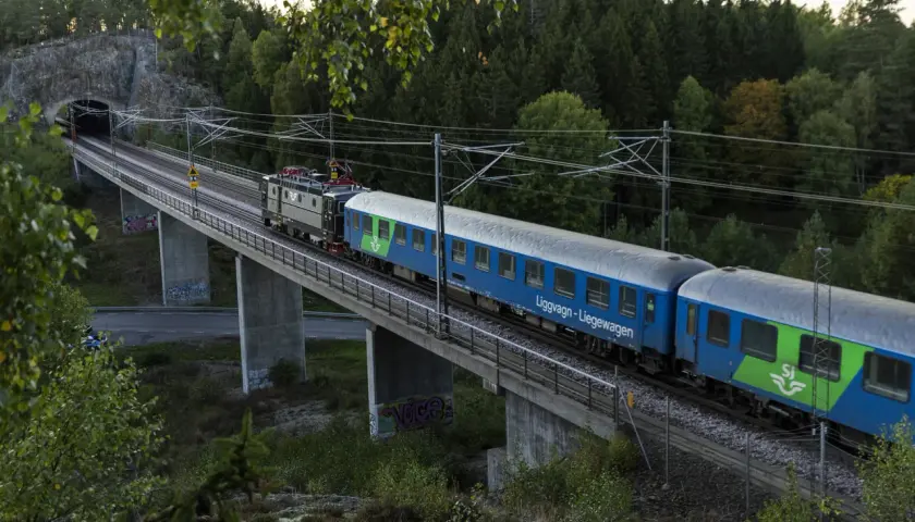 Ett blått SJ-tåg, kanske ett nattåg Berlin, korsar en bro genom en skogig miljö.