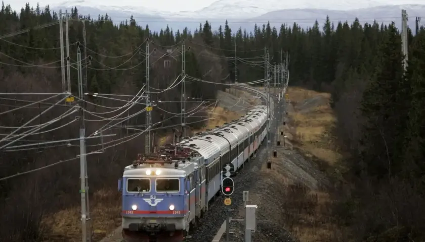 Ett SJ-tåg med nummer 1343 på väg mot Trondheim kurvar genom en skog med snöklädda berg i bakgrunden.