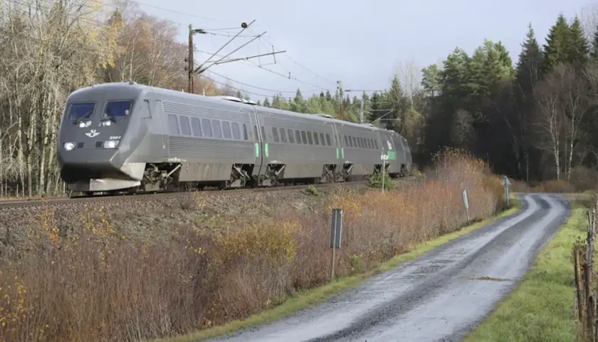Grått SJ-tåg på väg mot Stockholm, passerar en slingrande grusväg och höstfärgad natur.
