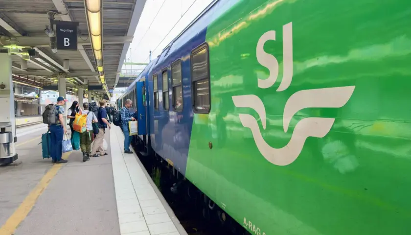 Folk väntar på ett grönt och blått tåg med SJ-loggan på perrongen. Kanske på väg till London?