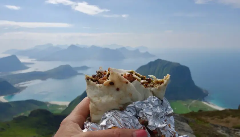 En hand håller en burrito med en fantastisk utsikt över Lofoten. En perfekt paus under vandringen.