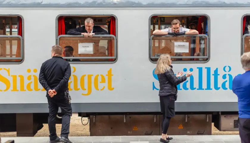 Män tittar ut genom fönstret på ett tåg med texten "Snälltåget" och destinationen Innsbruck. En kvinna gestikulerar mot tåget.