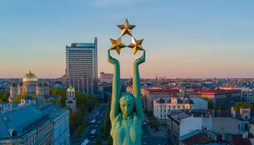 Frihetsmonumentet i Riga reser sig mot en solnedgångshimmel, med tre gyllene stjärnor högt upp. En båt syns i fjärran.