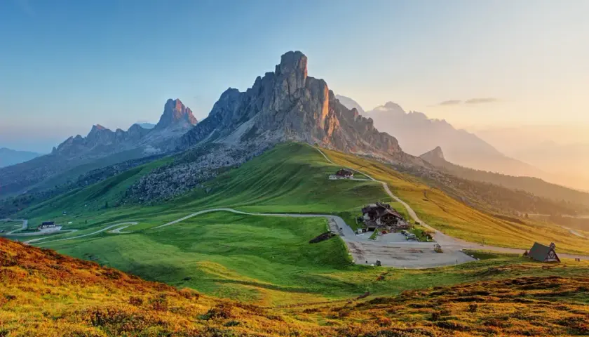 Gröna kullar och dramatiska bergstoppar i Dolomiterna sommar. En slingrande väg leder upp till ett hus vid foten av ett mäktigt berg.