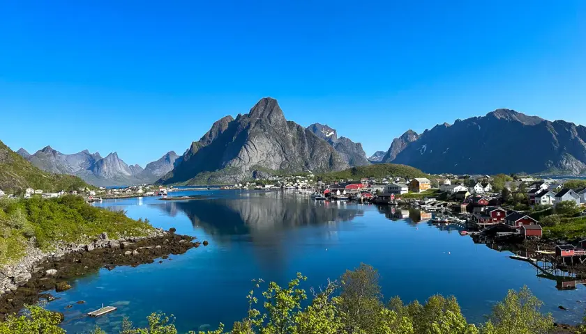 En klarblå himmel speglar sig i det lugna vattnet vid en idyllisk by. Majestätiska berg reser sig bakom de röda husen. En vacker lofoten sommarvy.