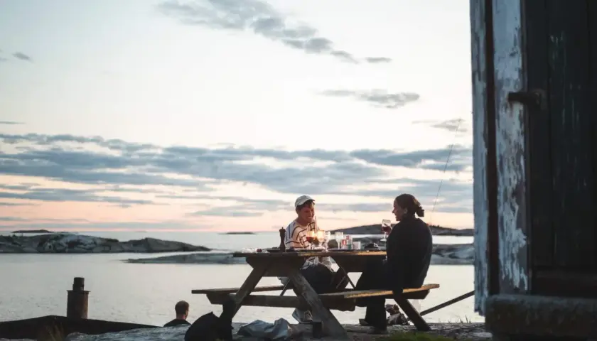 Två personer njuter av en middag vid ett picknickbord vid vattnet. En perfekt hemester i Stockholms skärgård.