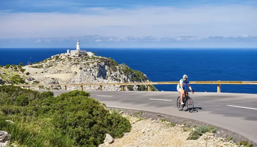 En cyklist njuter av en cykelsemester i Europa, med en fyr på en klippa vid det blå havet i bakgrunden.